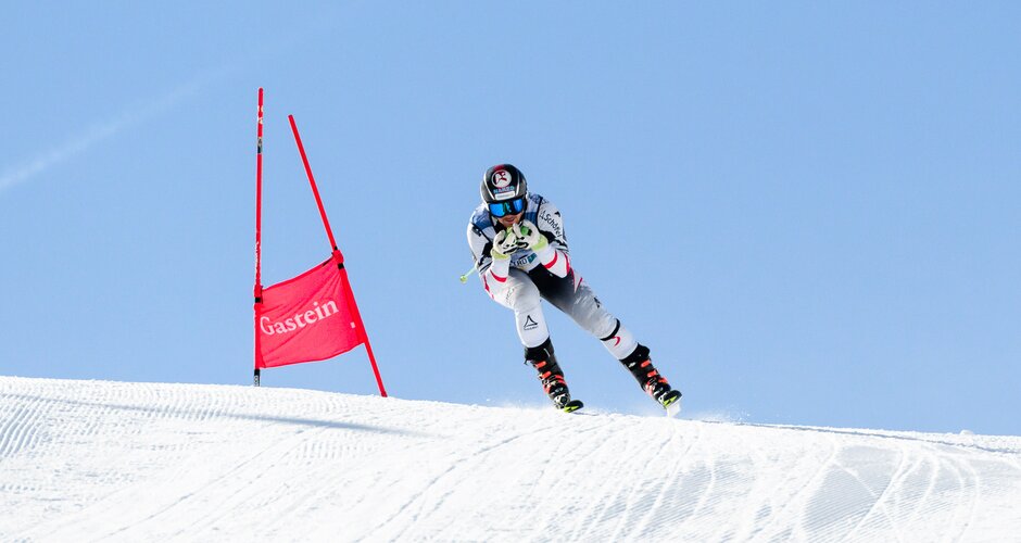 A ski racer in low tuck position descends a slope past a gate flag under blue sky. | © Gasteinertal Tourismus GmbH, Marktl Photograhpy
