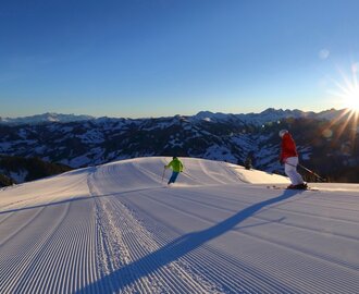 Two skiers on freshly groomed slope at sunrise with alpine mountain panorama