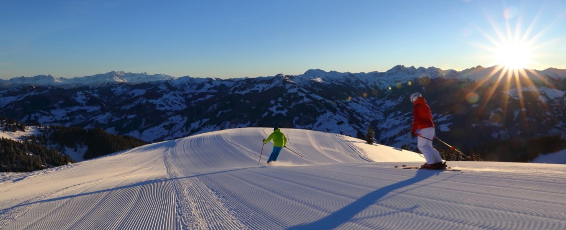 Two skiers on freshly groomed slope at sunrise with alpine mountain panorama