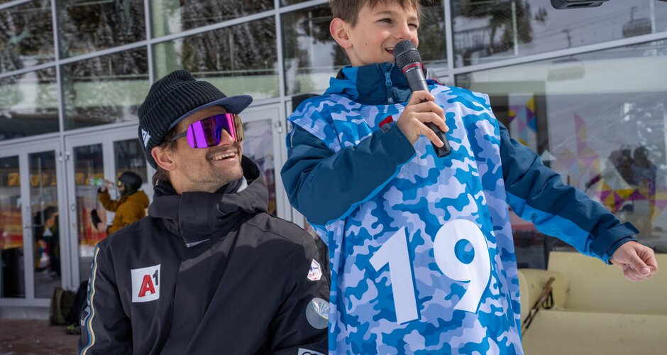 Young participant in blue jersey speaks into the microphone next to man in ski clothes