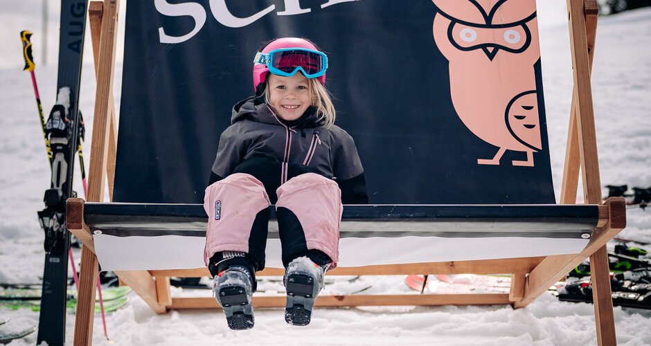 Smiling child in ski gear sitting in the snow on a large deck chair with Rose im Schnee banner
