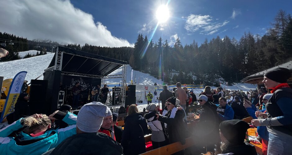 Many people in ski gear stand or sit on the terrace of the ski hut and look at a small stage on which musicians play. | © Hauser Kaibling