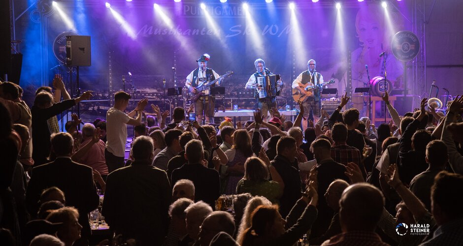 Three musicians with two guitars and a Styrian harmonica stand on the stage while many couples dance to them on the dance floor | © Harald Steiner 