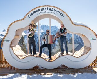Three musicians with tuba, accordion and guitar perform on wooden platform in snowy mountain scene | © Harald Steiner