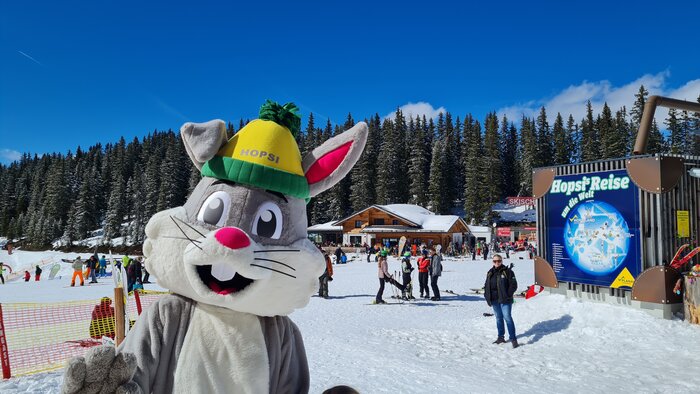 Hopsi the bunny mascot smiles at the camera and the family party takes place behind him | © Planai
