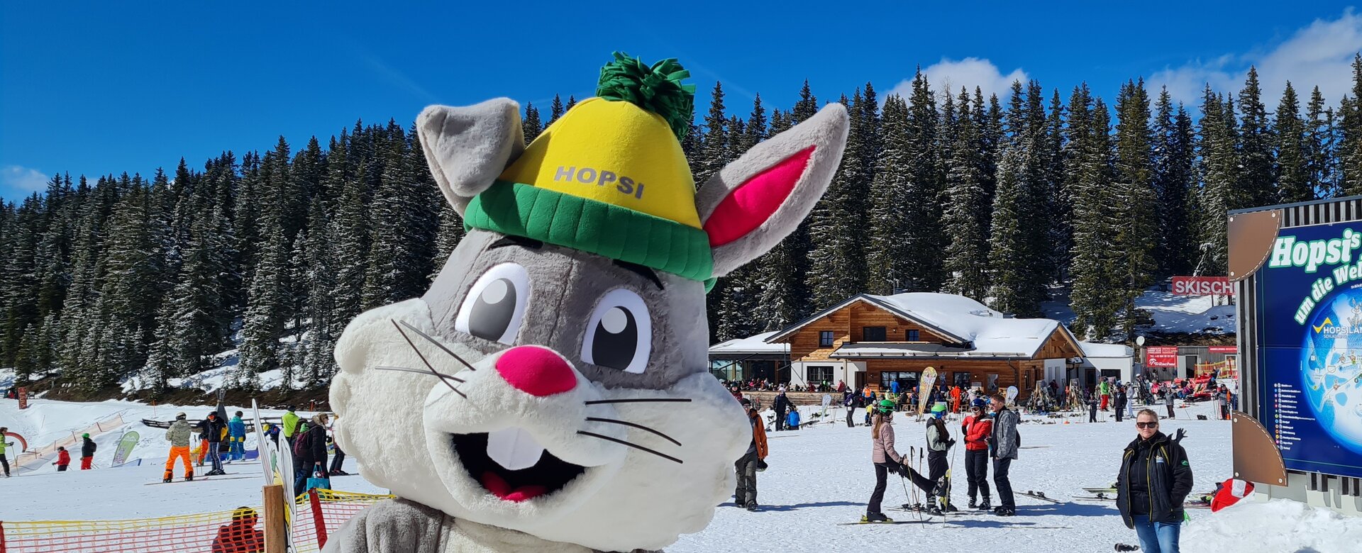 Hopsi the bunny mascot smiles at the camera and the family party takes place behind him | © Planai