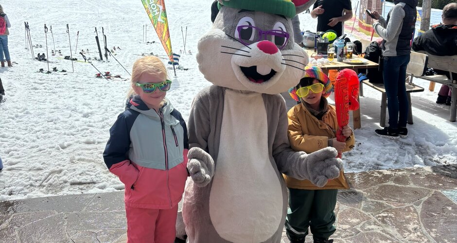 Two children stand with mascot Hopsi on a sunny terrace in the snow in front of ski slope and fores | © Planai