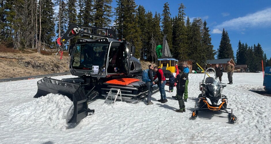 Several people stand on snow beside a snow groomer and a snowmobile in front of forest under sunny sky | © Planai