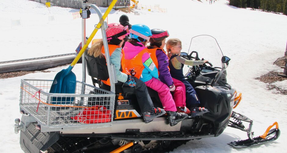 Four children wearing helmets and vests sit on a snowmobile on snow near a ski slope with forest | © Planai