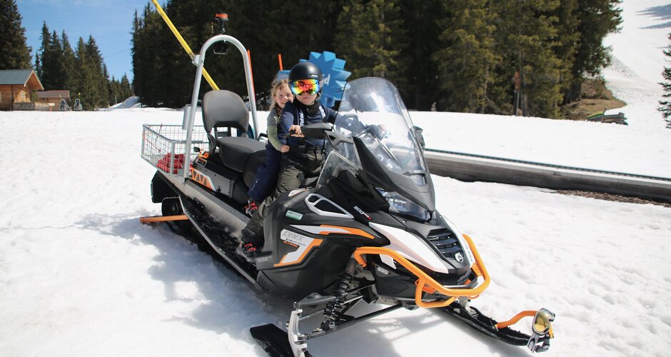 Two children wearing helmets sit on a snowmobile on snow with forest and ski slope in sunlight | © Planai
