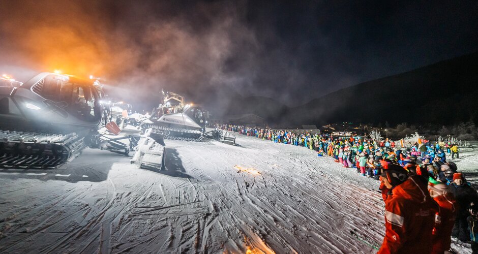 Illuminated snow groomers drive before large crowd in snow during nighttime New Year’s Eve show | © Gerald Grünwald