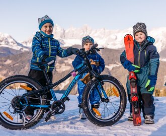 Three kids stand in snow, one with bike, one with skis, mountain panorama in the background | © Gerald Grünwald