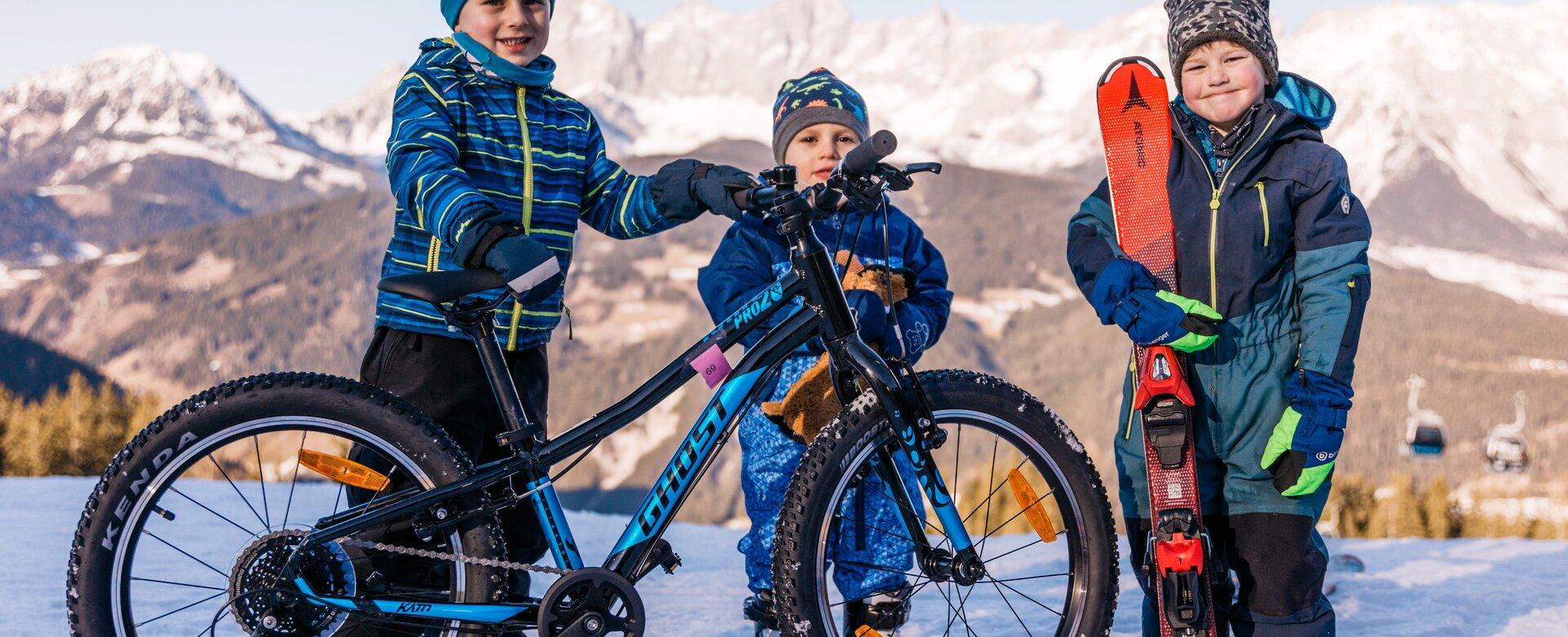 Three kids stand in snow, one with bike, one with skis, mountain panorama in the background | © Gerald Grünwald