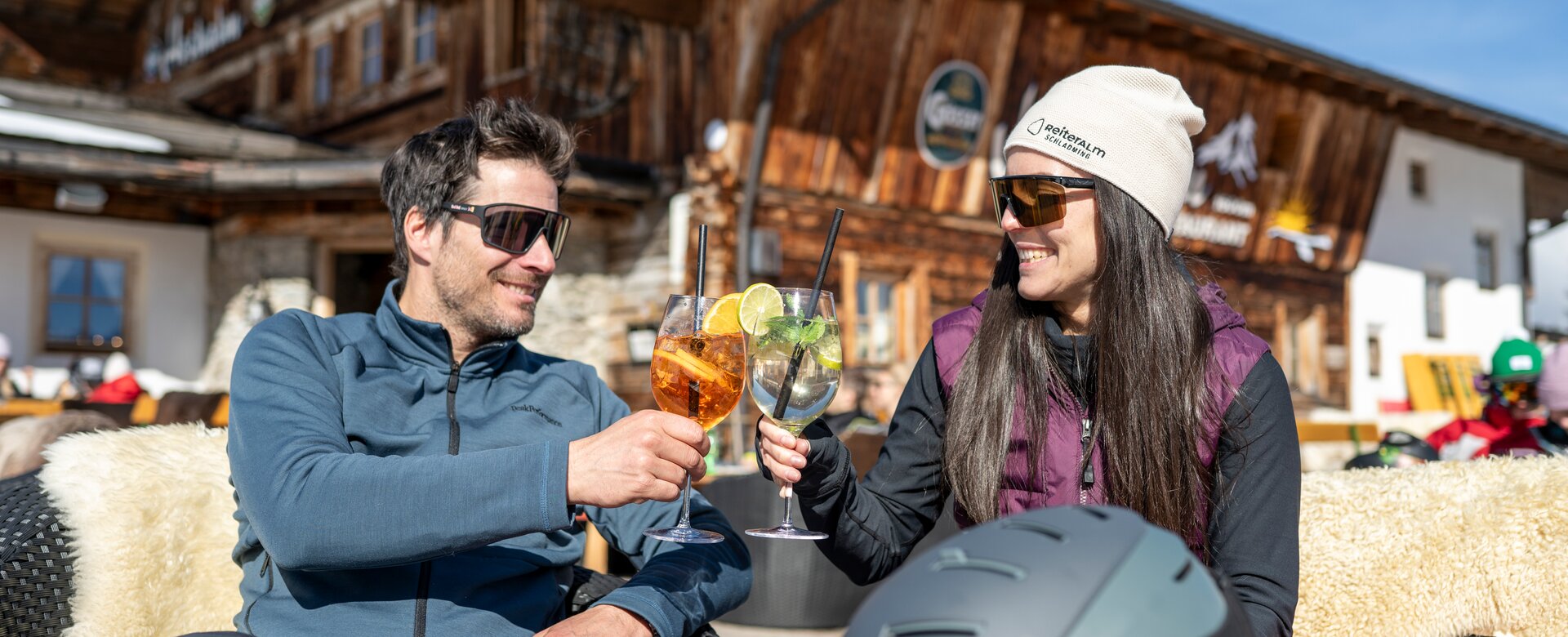 Man and woman toast with drinks in the sun, mountain hut and ski helmet in the background | © Lorenz Masser