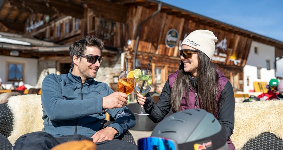 Man and woman toast with drinks in the sun, mountain hut and ski helmet in the background | © Lorenz Masser
