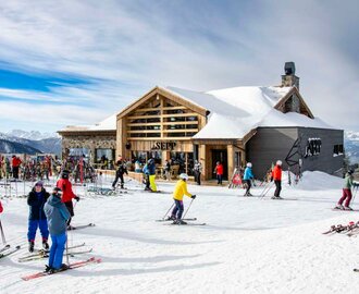 Exterior view of a modern ski hut with lots of skis out front and skiers passing by | © Christian Steiner