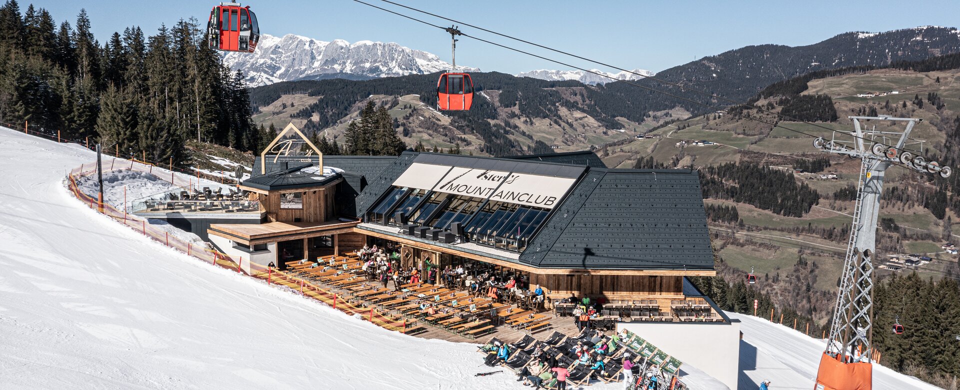Mountain lodge with sun terrace on a ski slope, red gondola cabins above and snow-covered peaks behind