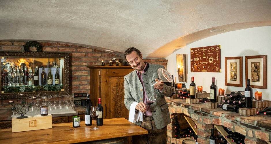 Host in traditional attire pours red wine from decanter in brick wine cellar