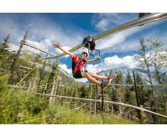 Young man rides the Flying Coaster through the forest on rails, arms wide open, mountains and blue sky in the background | © Christoph Huber