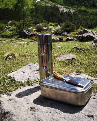 Metall-Jausenbox, Messer und Thermosflasche auf Fels in Almwiese platziert