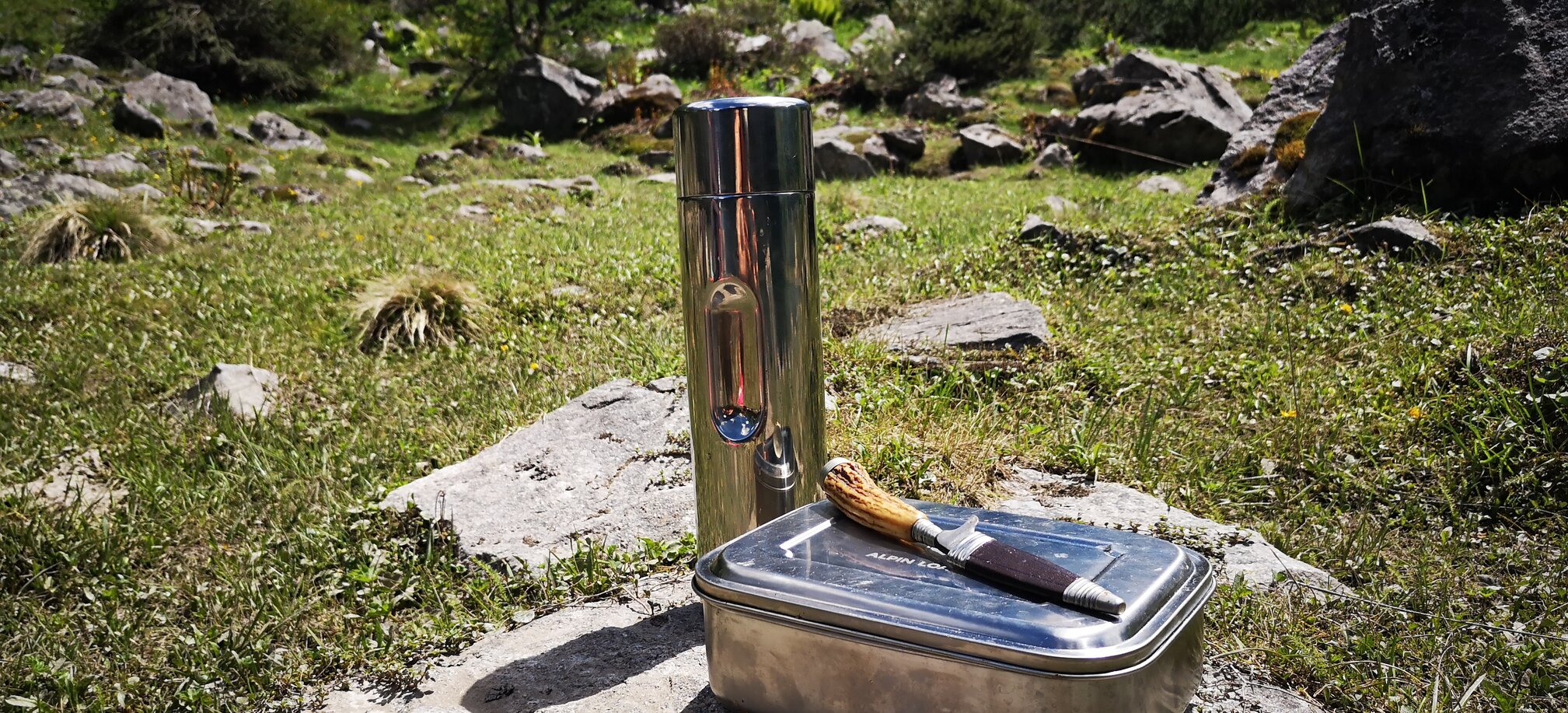 Metall-Jausenbox, Messer und Thermosflasche auf Fels in Almwiese platziert