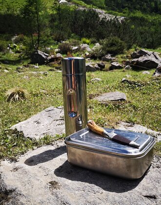 Metall-Jausenbox, Messer und Thermosflasche auf Fels in Almwiese platziert