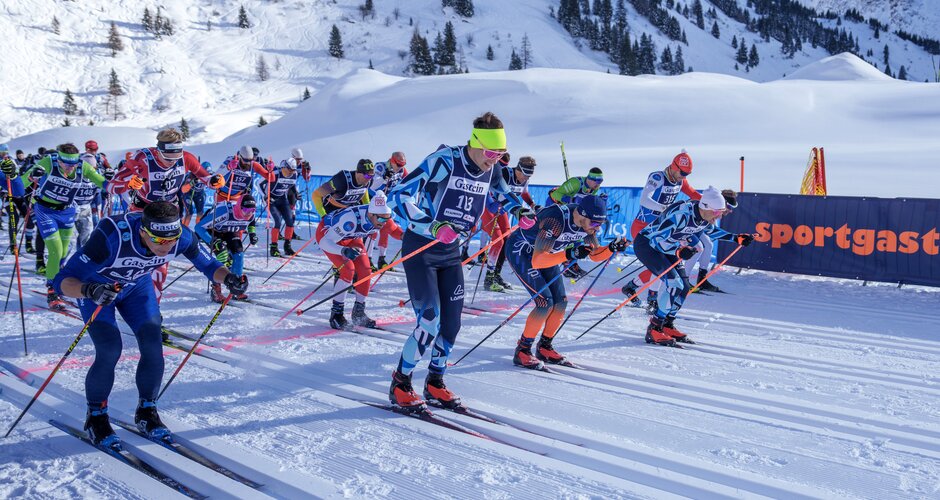 Numerous male cross-country skiers start side by side in bright sunlight on a groomed snowy track. | © Gasteinertal Tourismus, Christoph Oberschneider