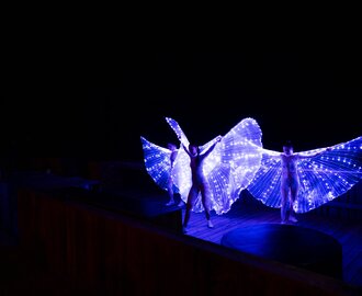 Three performers with LED wing costumes glowing in the dark on wooden terrace of mountain hut