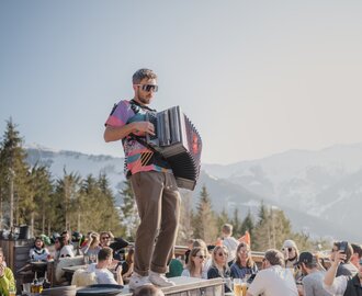Man with accordion plays on table among cheerful guests on sunny terrace with mountain view