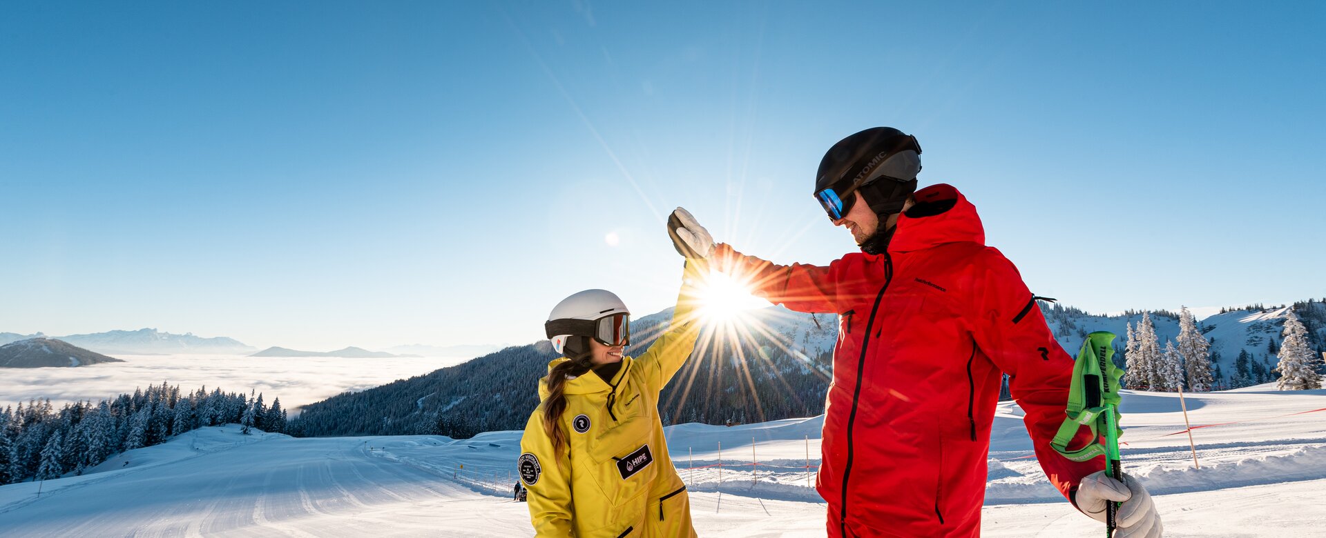 Two skiers in colourful suits give a high five on sunny, freshly groomed ski slope