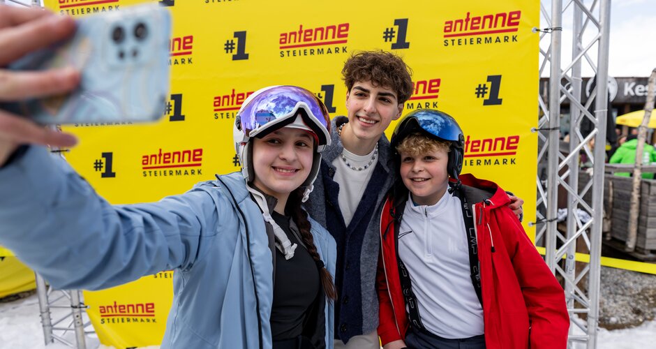 Three teenagers pose for a selfie in front of yellow Antenne Steiermark photo wall in snow. | © Ripix