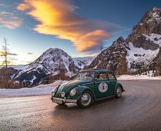 A green VW Beetle drives at sunset on snowy mountain road with alpine peaks in the background | © Ondrey Kroutil