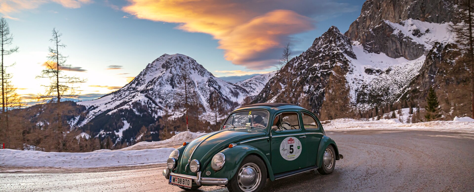 A green VW Beetle drives at sunset on snowy mountain road with alpine peaks in the background | © Ondrey Kroutil