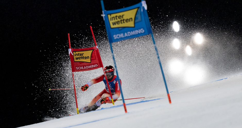 Alpine skier passes giant slalom gates at Schladming Nightrace, snow spray illuminated by bright floodlights. | © Martin Huber