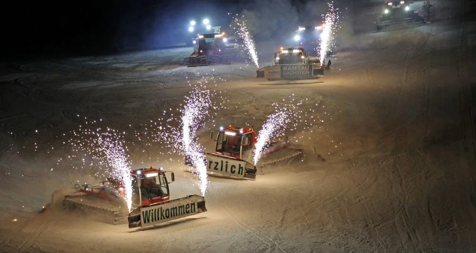 Piste machines descend one after the other on a floodlit piste at night in a formation | © Erlebnis Rittisberg