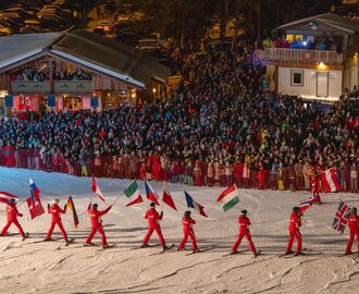 Skiers in red carry national flags on a slope at night in front of a large crowd | © Michael Simonlehner