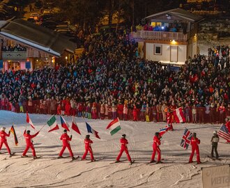Skiers in red carry national flags on a slope at night in front of a large crowd | © Michael Simonlehner