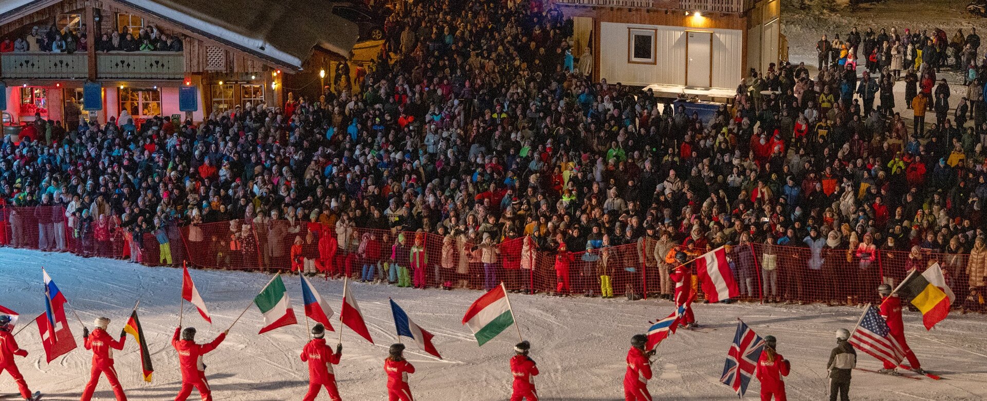 Skiers in red carry national flags on a slope at night in front of a large crowd | © Michael Simonlehner