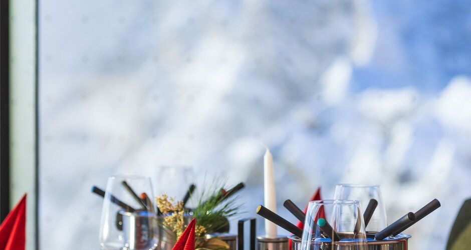 Set table with fondue, glasses and red napkins in front of window with snowy mountain view | © Martin Huber