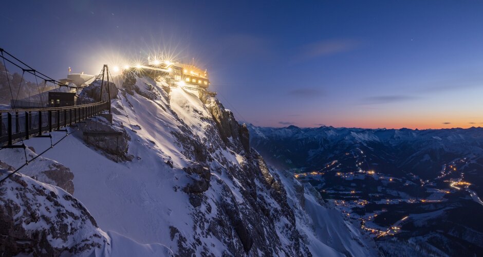 Illuminated mountain station with suspension bridge at dusk on Dachstein, valley lights below | © Martin Huber