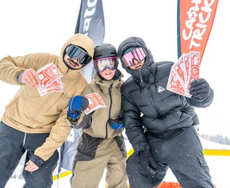 Three snowboarders hold red prize bills in front of flags during snowfall at Superpark Planai | © Jordan Sitzwohl