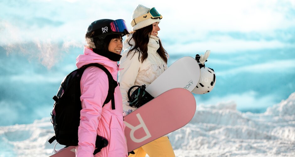 Two young ladies with snowboard equipment walk past the photographer on the piste and to the side | © QParks