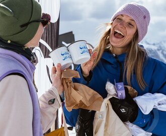 Two women toast with white enamel mugs, laughing in front of snowy mountain scenery. | © Henrie Kelbling