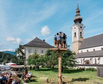 Men in traditional dress stand on a climbing pole at a craft festival in Altenmarkt, audience watches from benches outside. | © Matthias Fritzenwallner