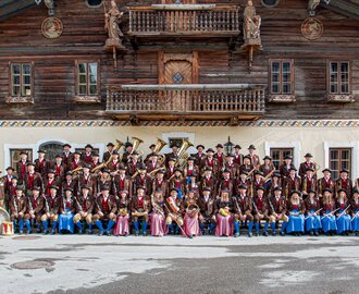 The Altenmarkt traditional band poses in costume with instruments in front of a rustic wooden house | © Trachtenmusikkapelle Altenmarkt