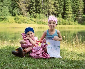 Two kids at the See.Musik event by the lake, sitting on grass with picnic bags and a self-drawn picture of sun and mountains. | © Matthias Fritzenwallner