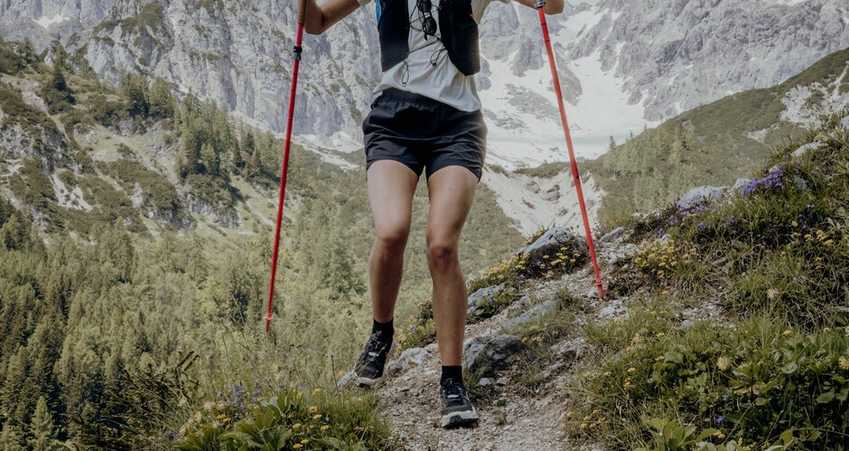 A female trail runner descends a rocky alpine path with poles, with the snow-covered Bischofsmütze peak in the background. | © ©CoenWeesjes_www.filzmoos.at