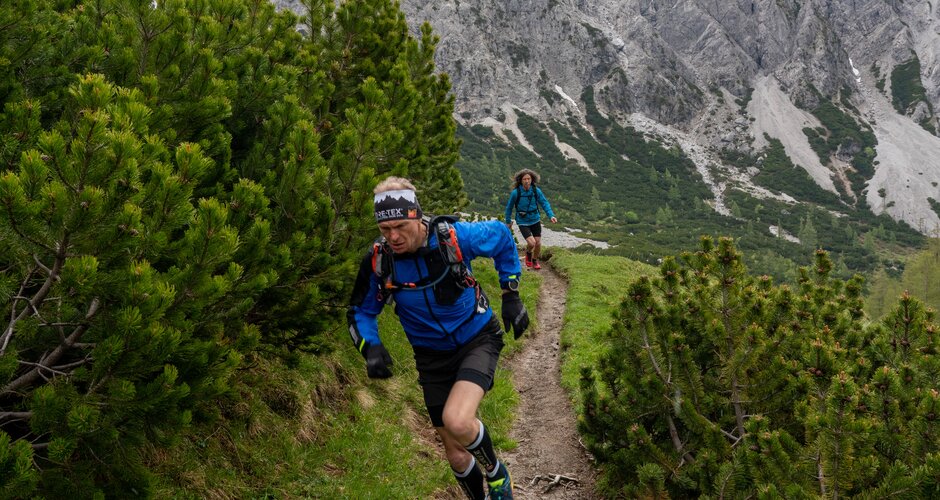 A trail runner climbs a narrow, steep mountain path through pines with snow-covered peaks towering in the background. | © ©CoenWeesjes_www.filzmoos.at