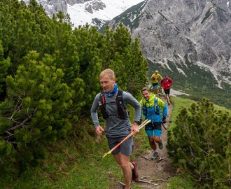 A group of trail runners moves along a narrow mountain path lined with pine bushes below snow-covered rock peaks. | © ©CoenWeesjes_www.filzmoos.at