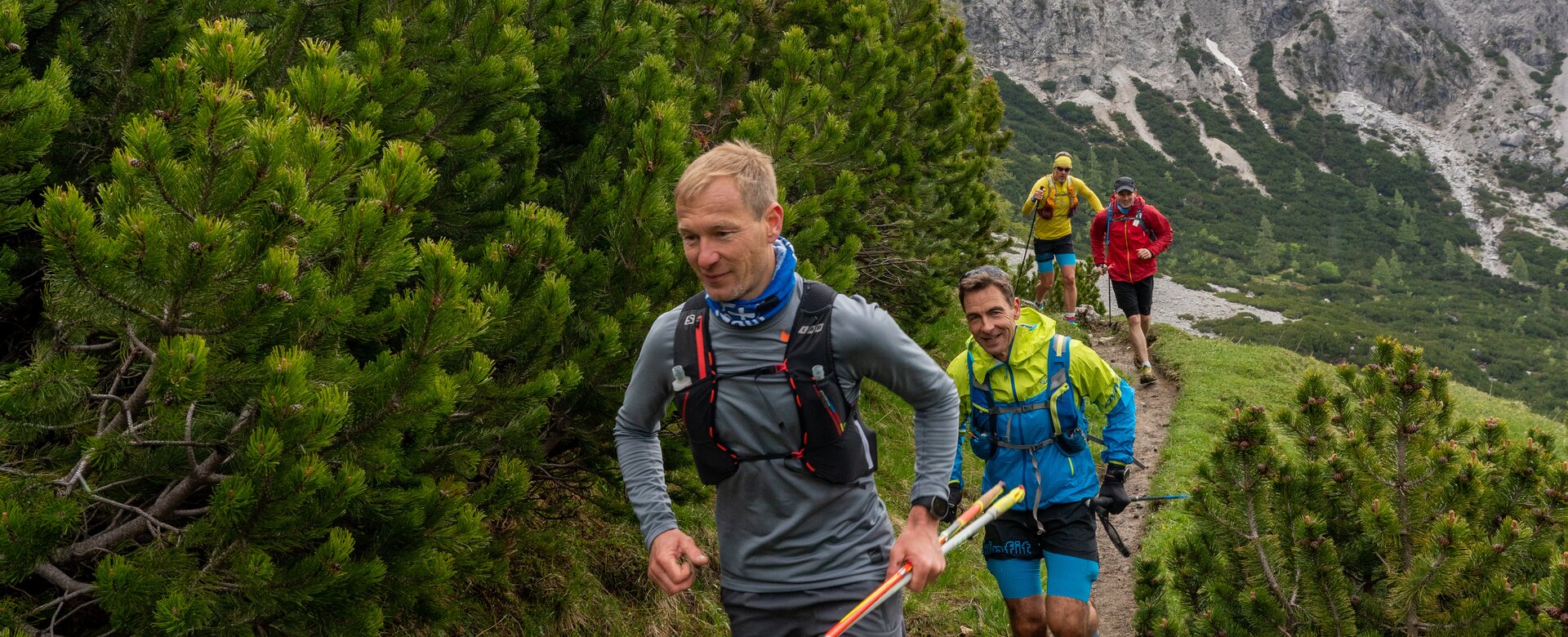 A group of trail runners moves along a narrow mountain path lined with pine bushes below snow-covered rock peaks. | © ©CoenWeesjes_www.filzmoos.at